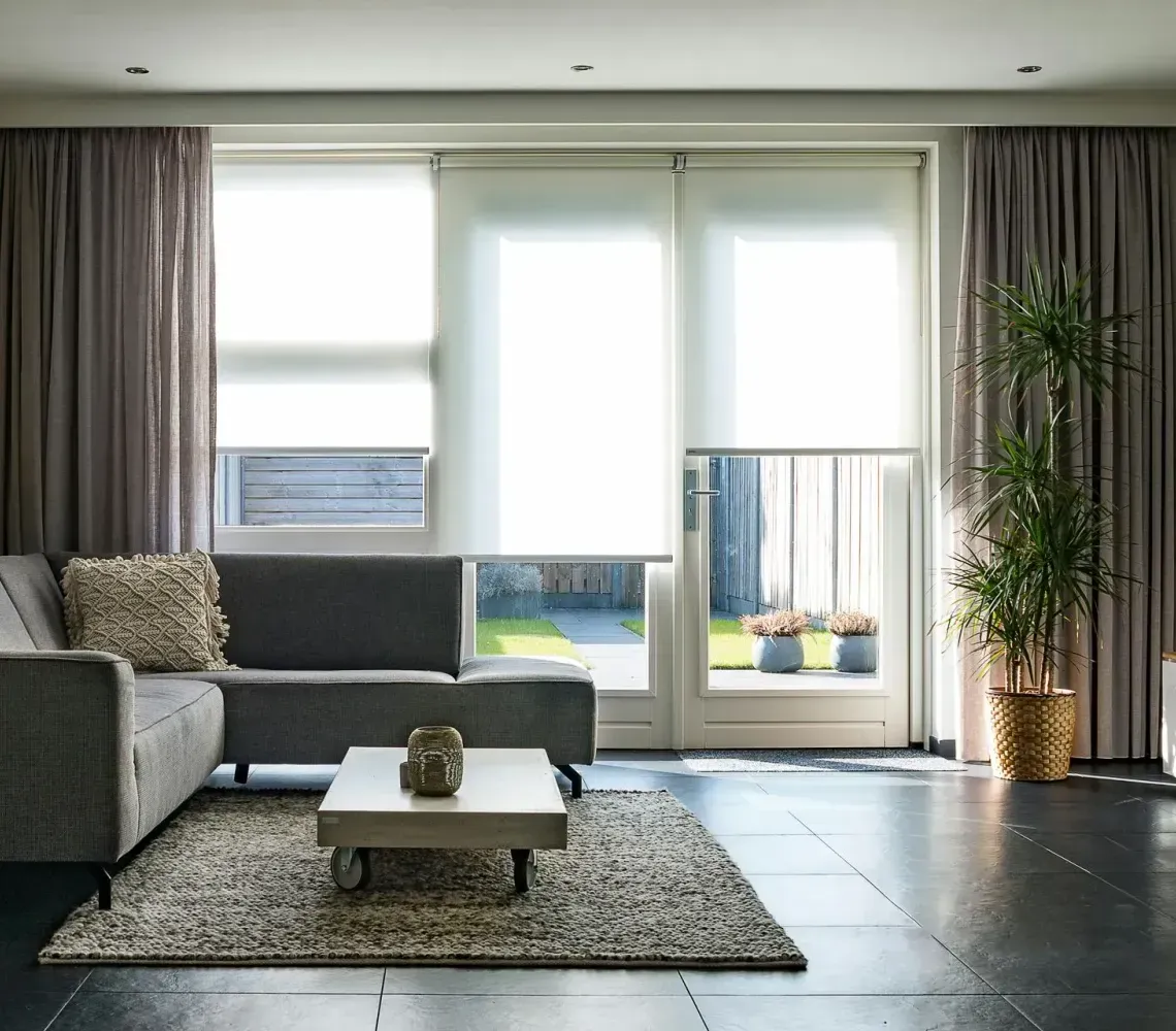 Modern living room with a gray sectional sofa, patterned area rug, coffee table, and large windows with light shades.