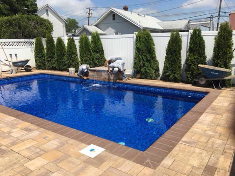 A fenced in pool with a brown brick patio that has 3 men working on the pool fountain