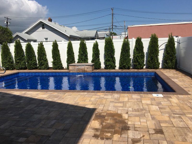 A fenced in ground pool with a brown brick patio surrounded by a white fence and green tall bushes