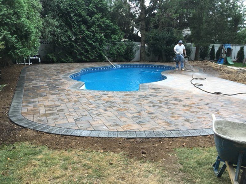 A in ground pool with grey bricks for patio while a man power washes the patio