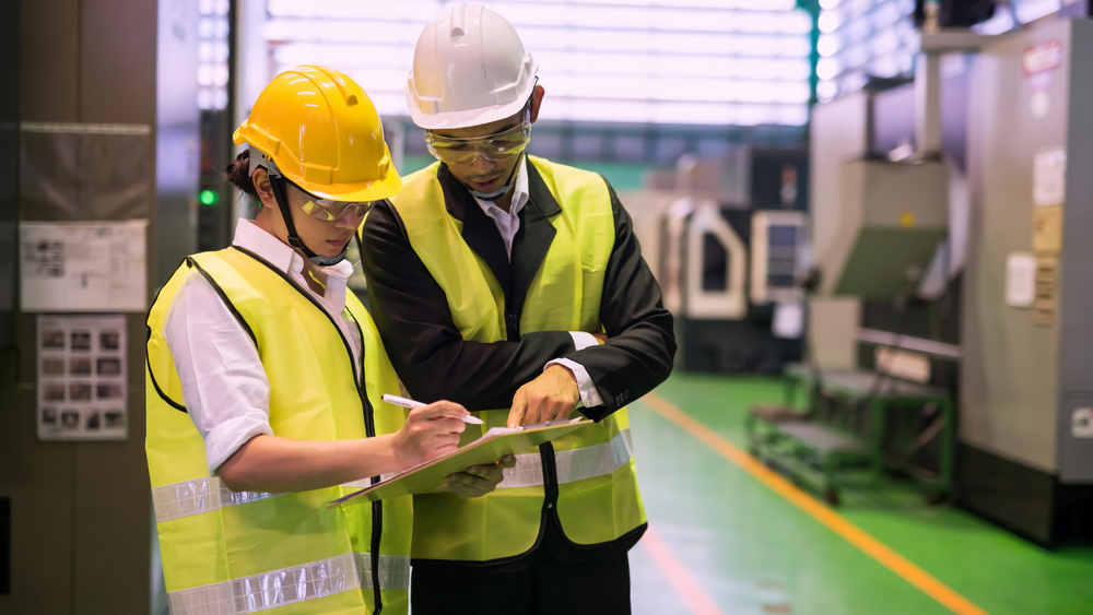 Two people in safety vests and hard hats review a clipboard in a factory.