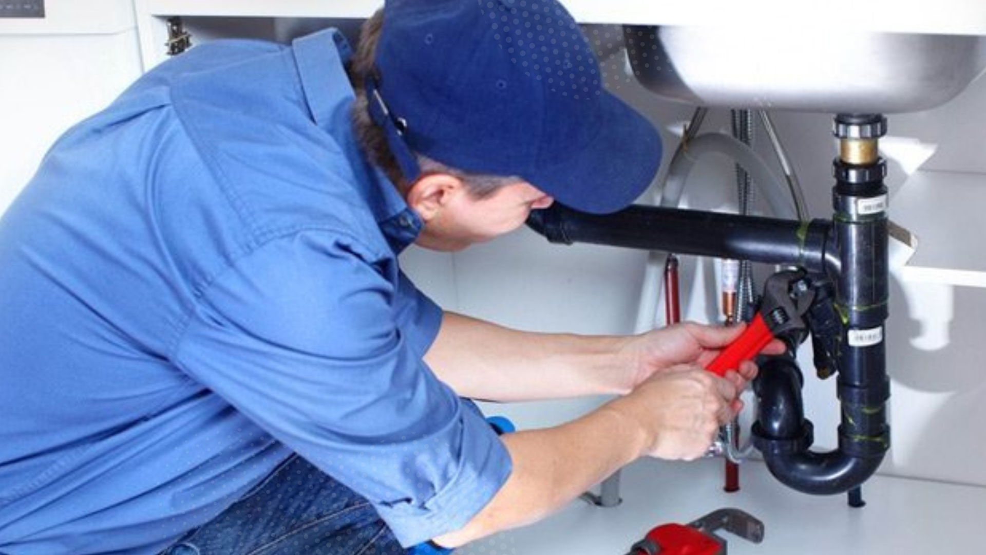 Plumber in blue shirt and cap using a wrench under a kitchen sink.