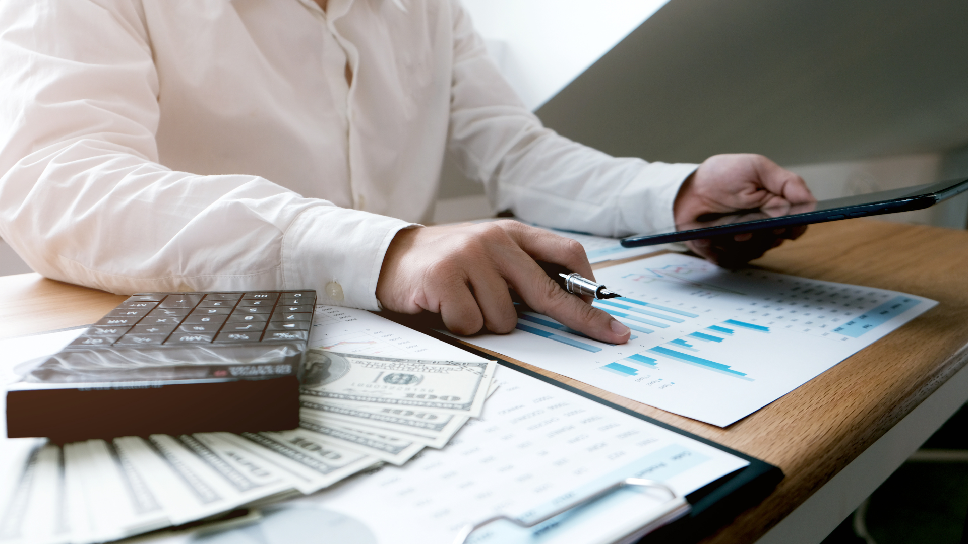 Person in white shirt analyzing financial graphs, pointing pen, with calculator and cash on desk.