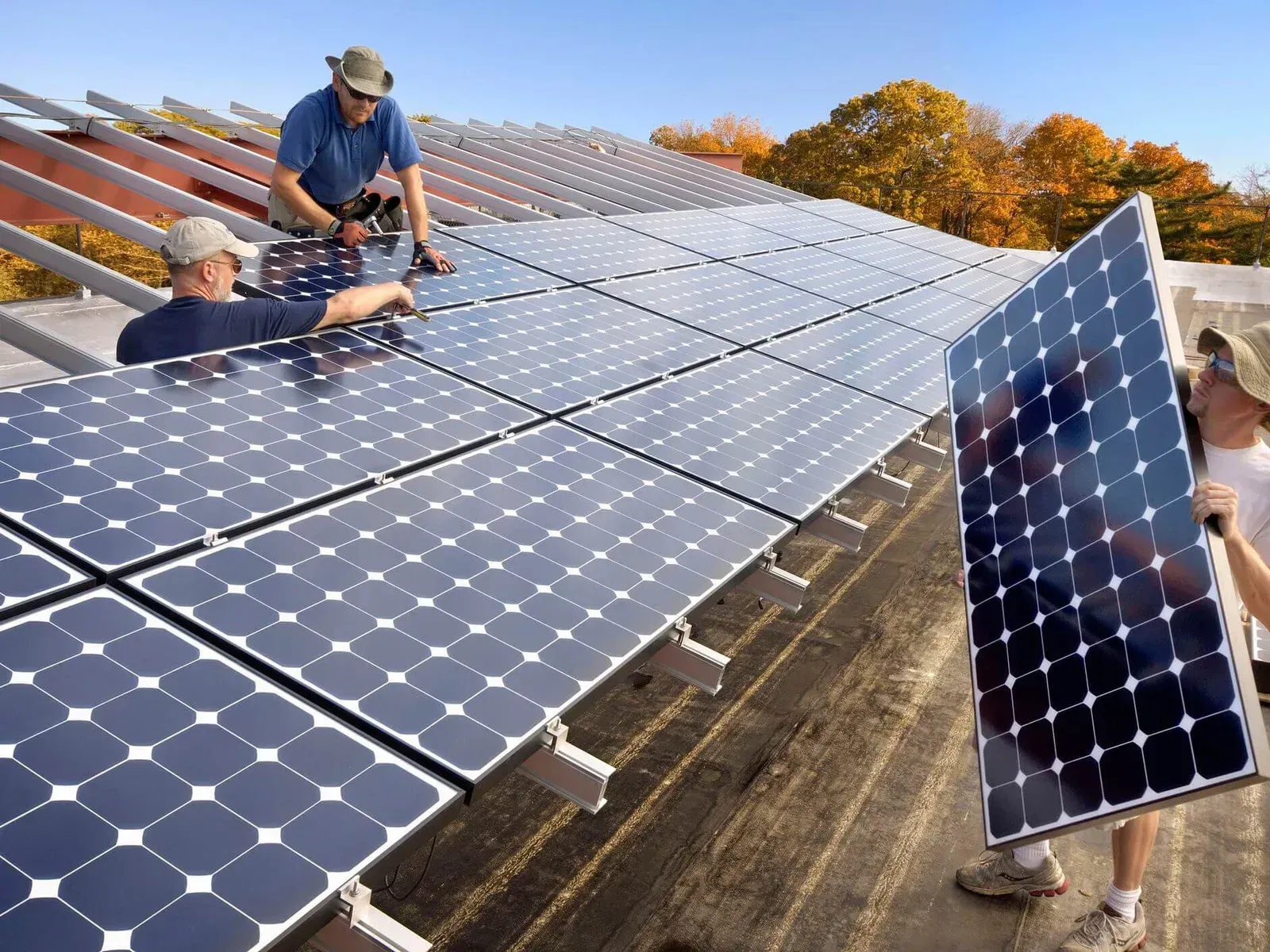 Workers installing solar panels on a roof. Blue panels against a blue sky, fall foliage in the background.