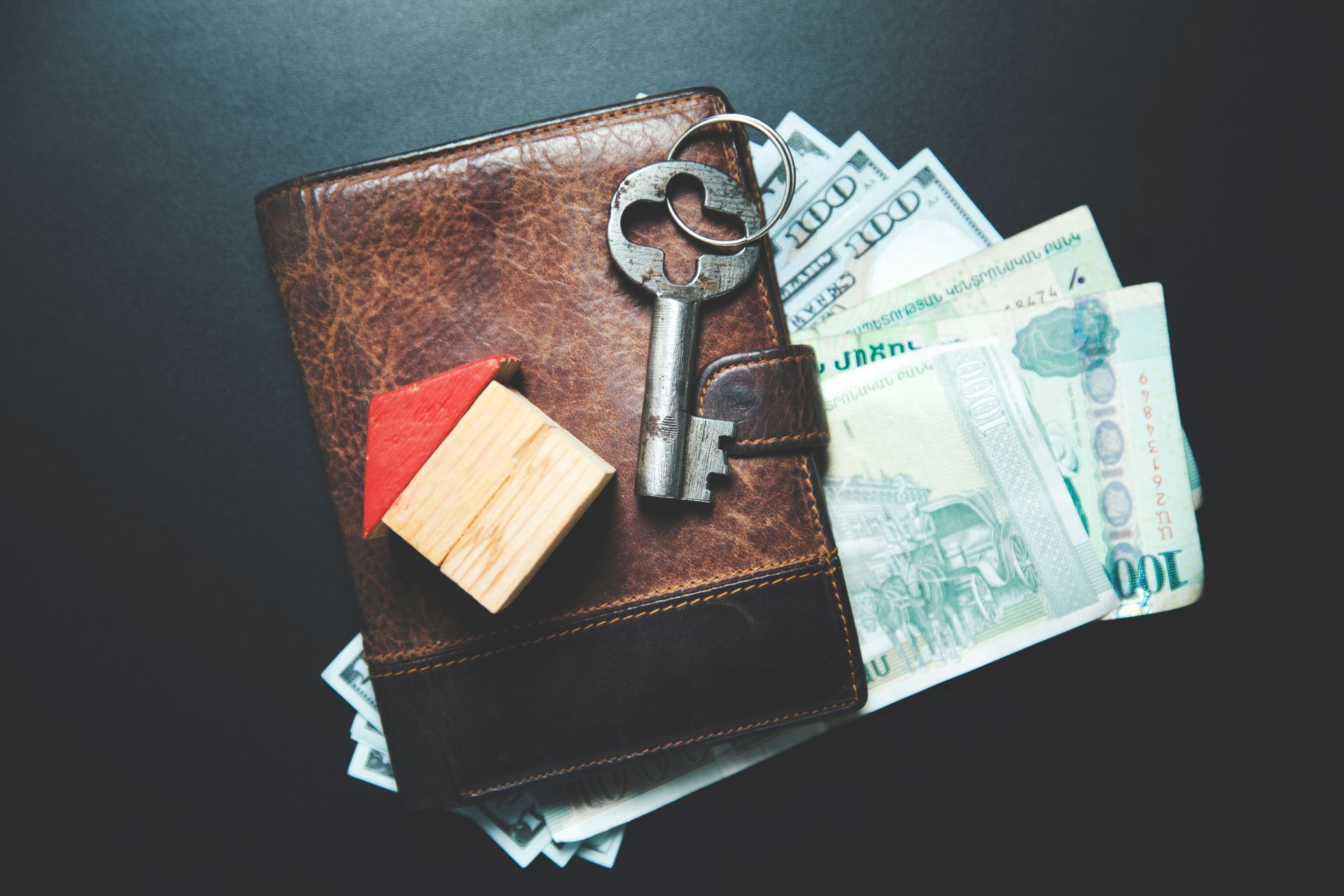 Brown wallet, key, and toy house with money on a black surface.