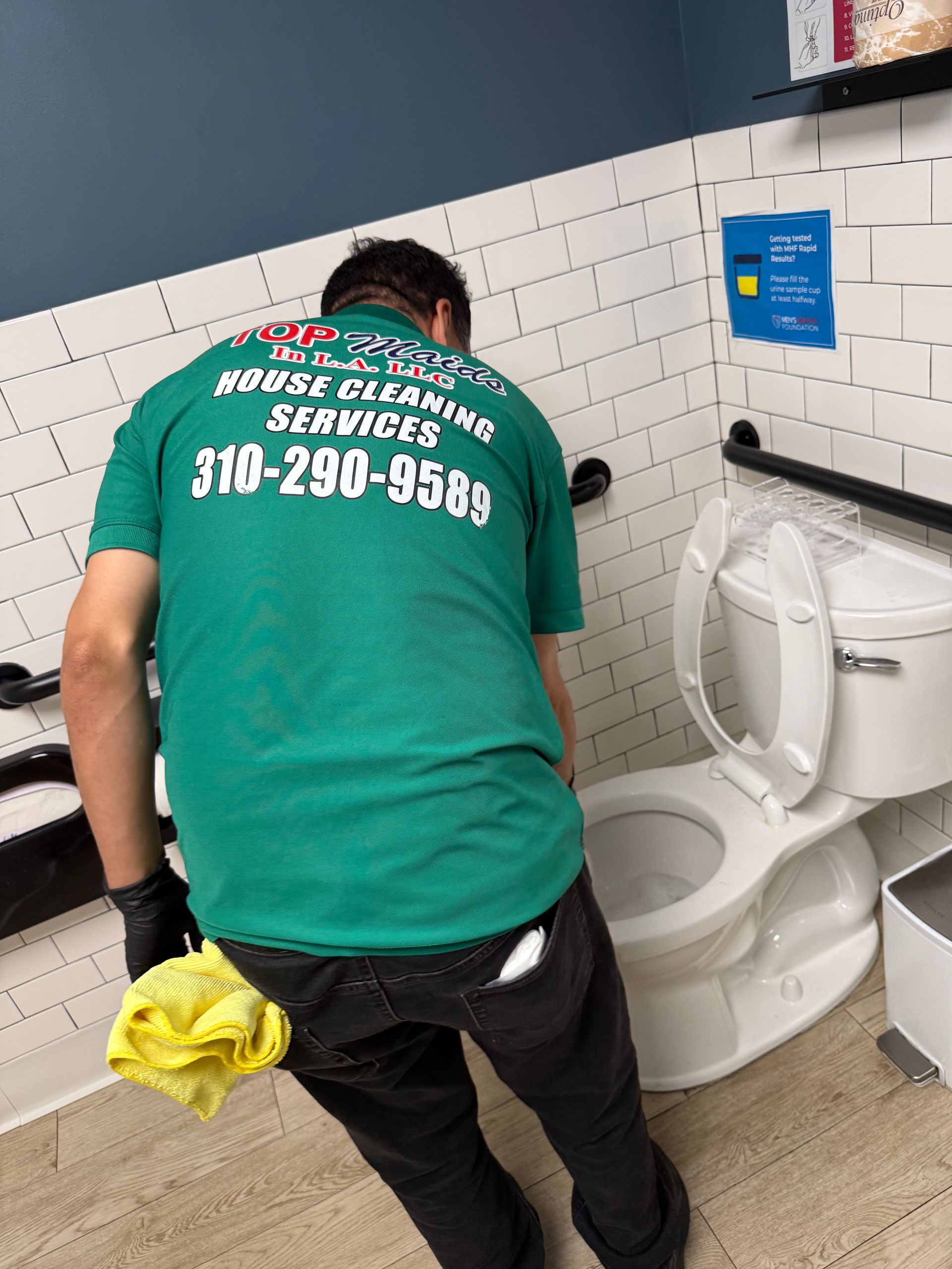 Person in green shirt cleaning a white toilet in a bathroom with a phone number on the shirt.