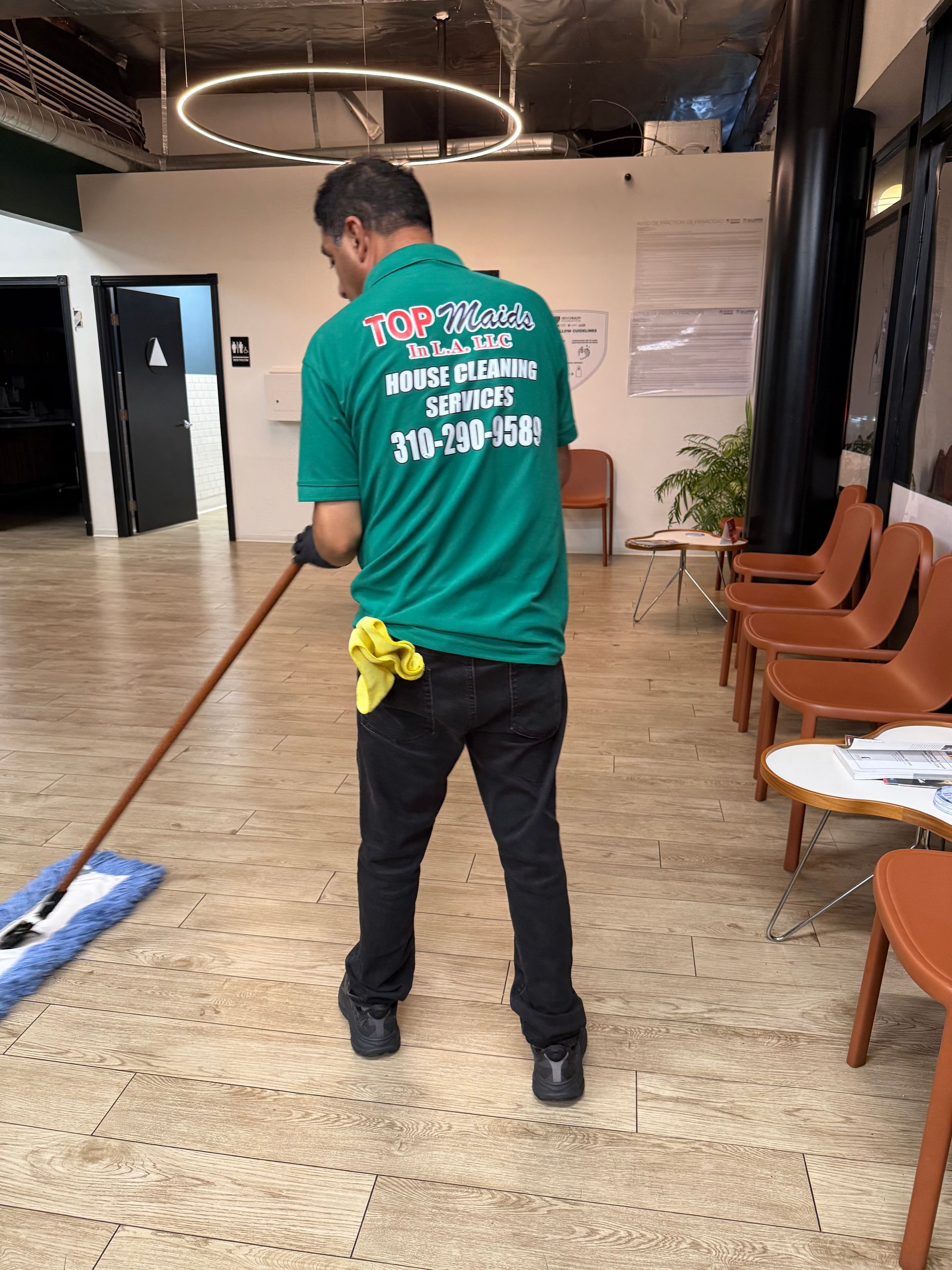 Man in green shirt mops floor in office. Brown chairs line the right, black column on the right.