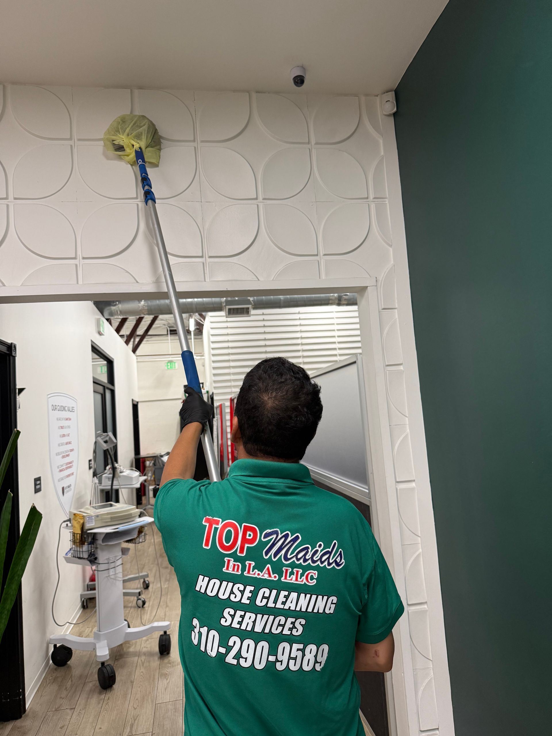 Person in green shirt cleaning ceiling with a long-handled mop. Indoors, white walls, doorway.