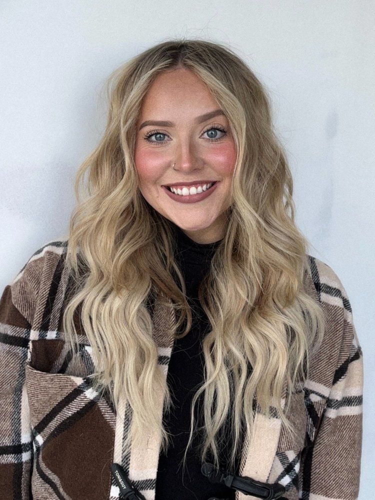 A woman with long blonde hair is wearing a plaid jacket and smiling.