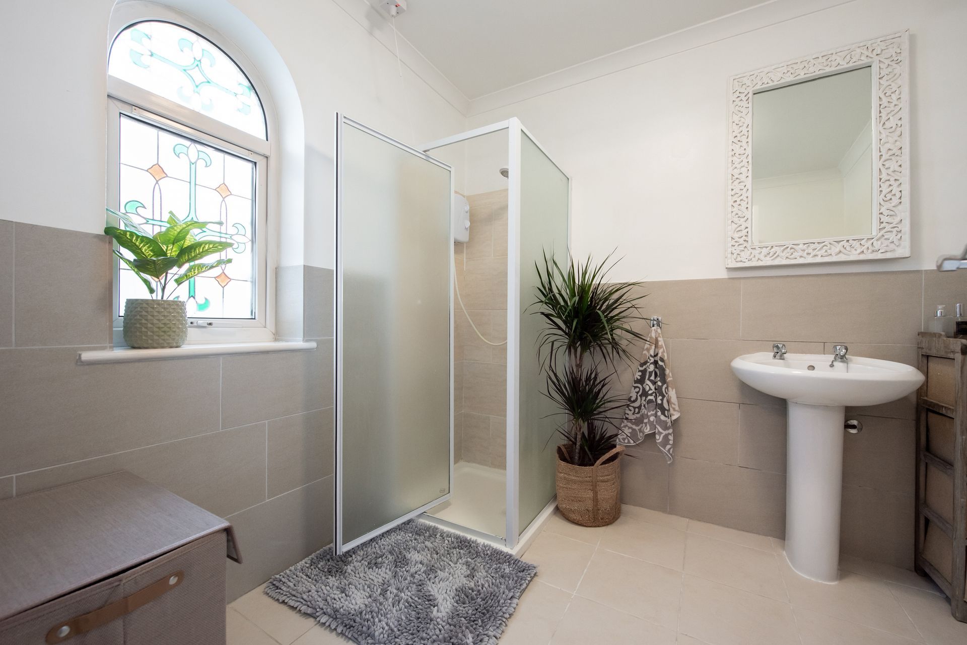 Bathroom with frosted glass shower, sink, window with plant, and decorative mirror. Neutral tones.