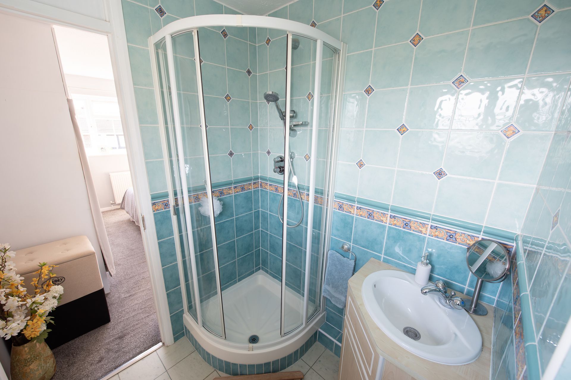 Small bathroom with teal tiled walls and a curved glass shower.  A small sink and the doorway to another room are also visible.