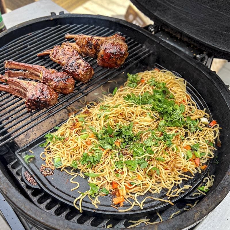 a skillet filled with noodles and lamb chop on a grill