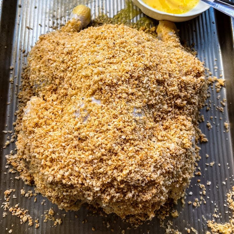 A whole brined chicken is covered in crumbs on a pan