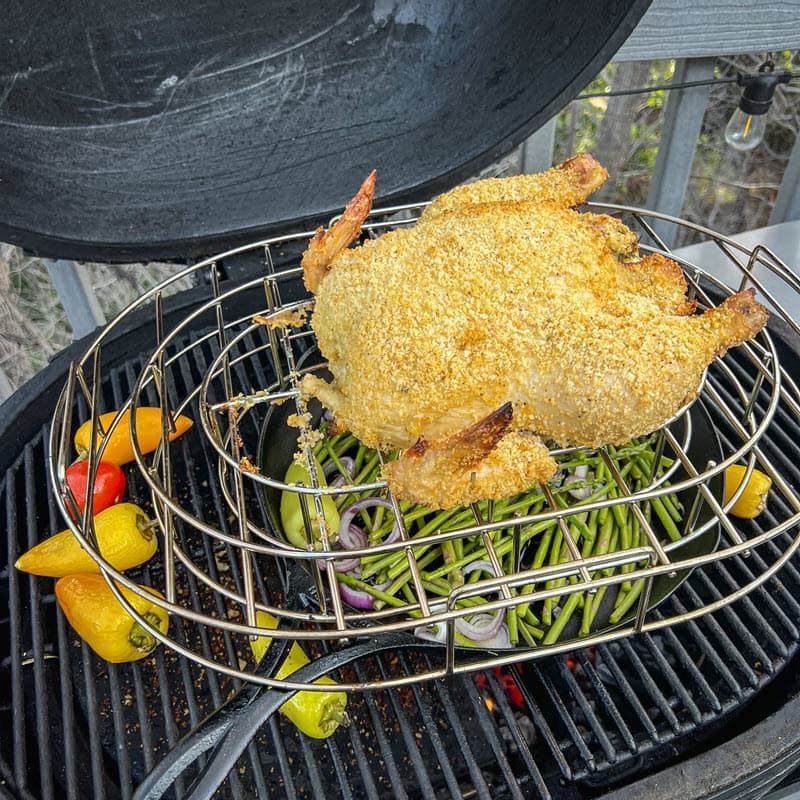 A whole panko-crusted chicken and peppers are cooking on a grill with green beans in a cast iron pan