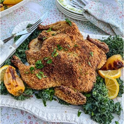 A whole grilled chicken with crust is sitting on top of a white plate with lemons and garnish.