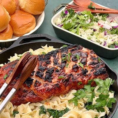 a skillet filled with noodles , pork loin and tongs with rolls and salad on a table