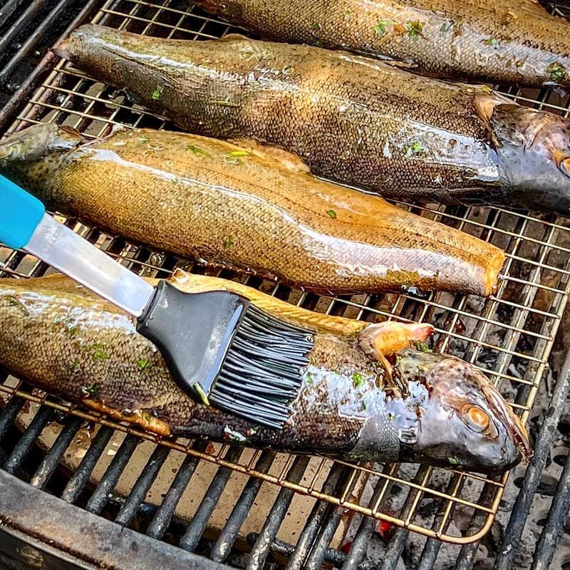 A bunch of fish are being cooked on a grill and marinated with oil on a brush.