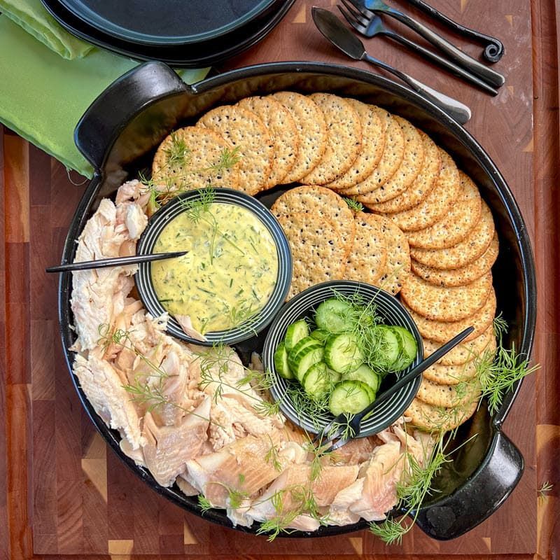 A tray of crackers cucumbers and fish with a mustard dill sauce on a wooden table