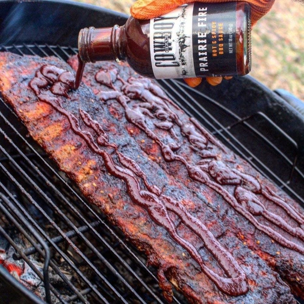hand pouring Cowboy Prairie Fire BBQ Sauce over ribs cooking on grill