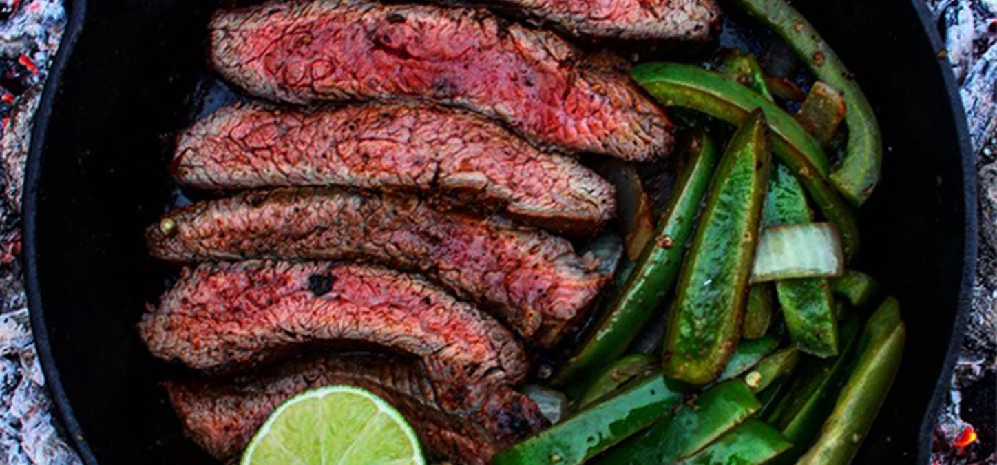 Flank steak, green bell peppers and a lime cooking over live coals in a cast iron pan