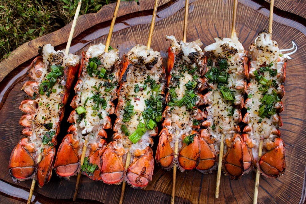Grilled lobster tails on skewers with seasoning. The lobster skewers are on wooden platter