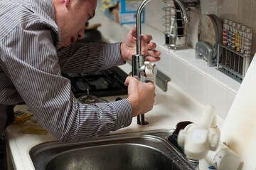 Picture of a Handyman wearing a striped long sleeve button up shirt repairing a sink faucet.