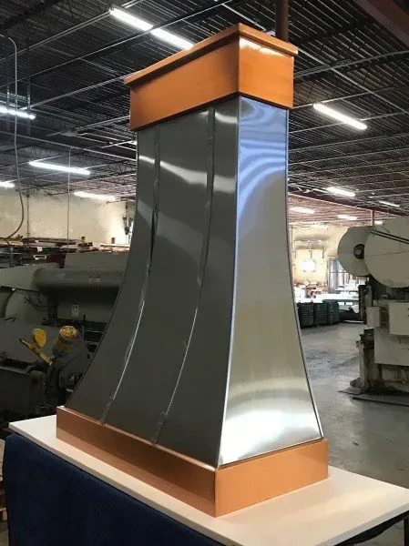 A stainless steel and copper hood is sitting on a table in a factory.