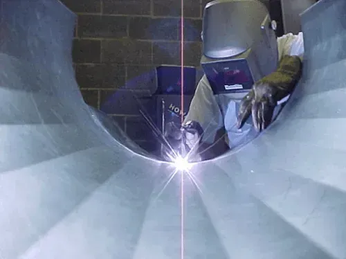 A man wearing a welding helmet is welding a metal pipe.