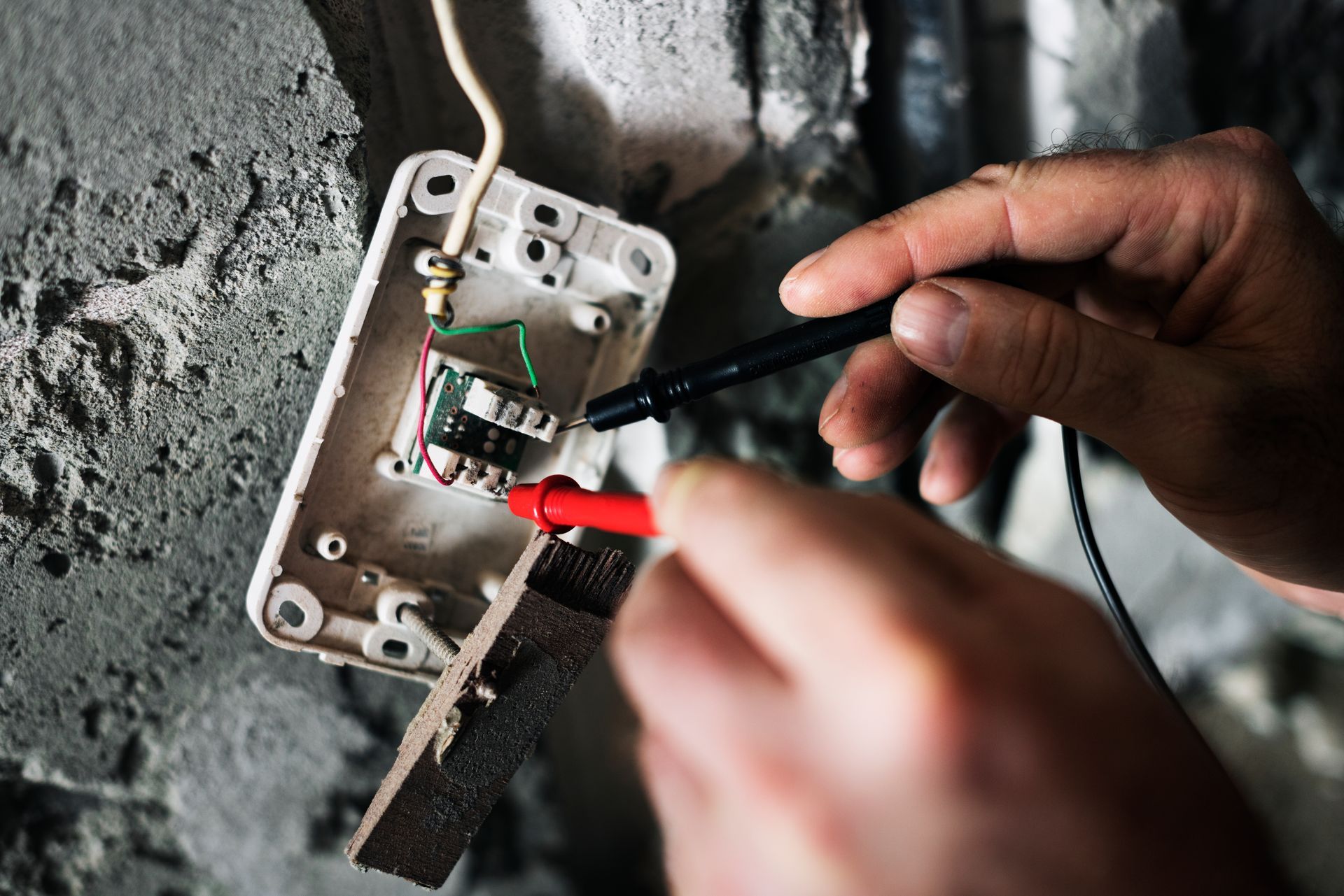 Hands testing electrical wiring with red and black probes, in a gray wall opening.