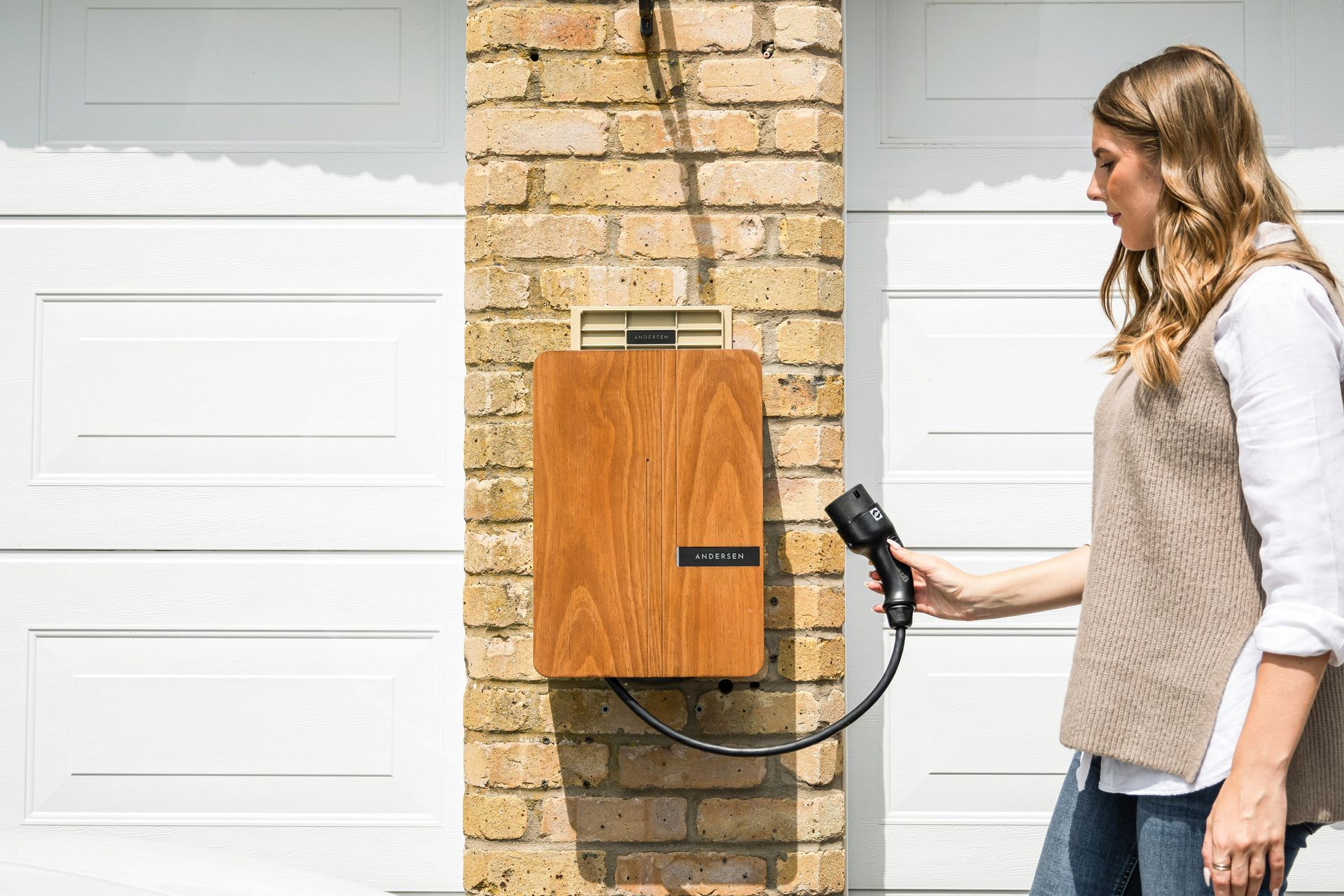 Woman plugs in an EV charger on a brick wall next to a garage door.