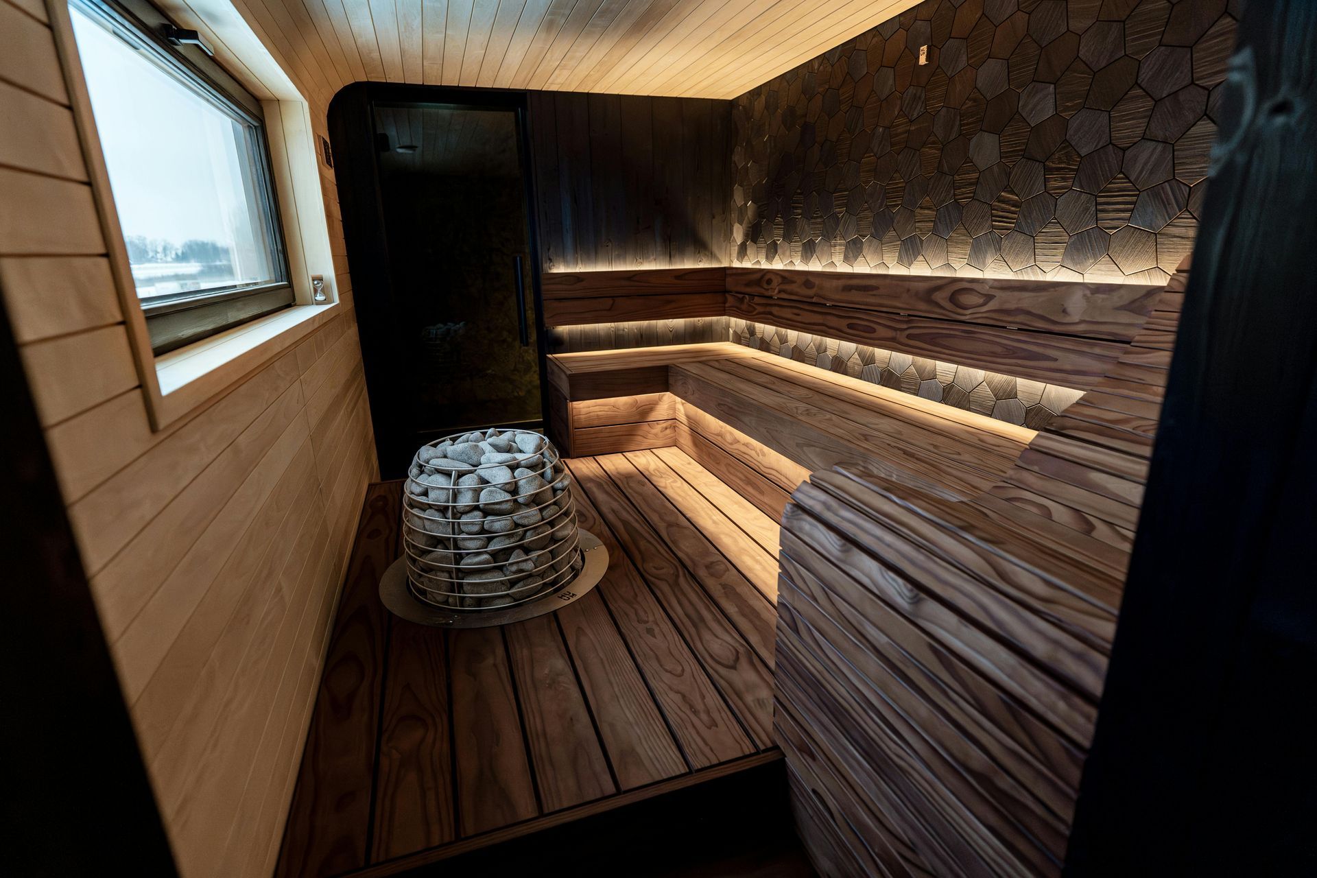 Wooden sauna interior with stone-covered heater and window, illuminated by warm lighting.