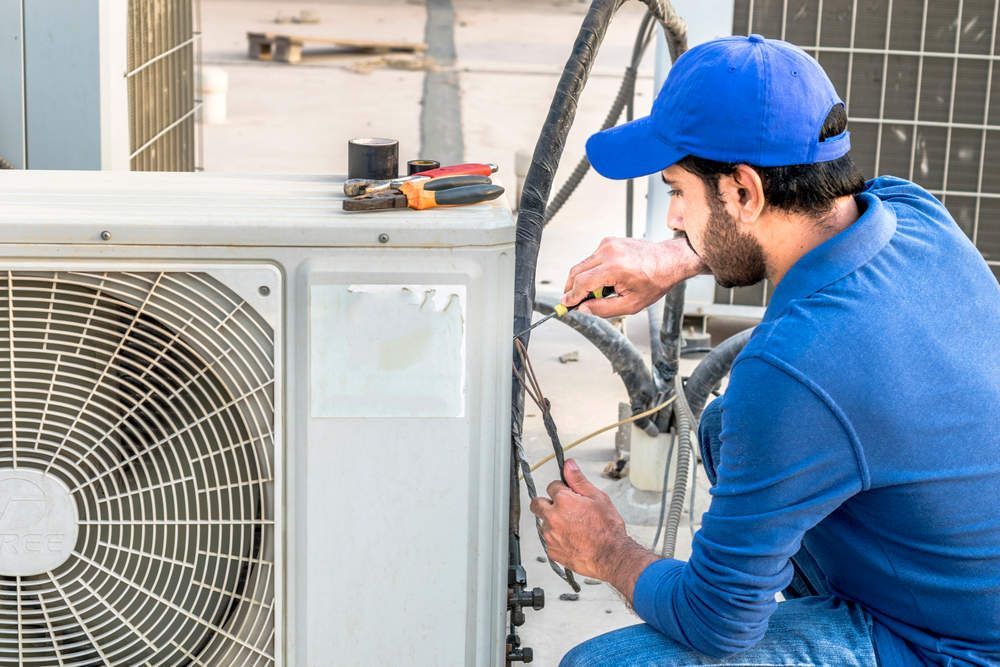 A person in a blue shirt and cap repairs an outdoor air conditioning unit on a rooftop.
