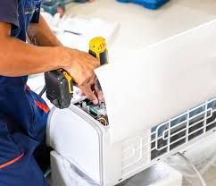 A technician using a cordless drill to repair or install a white wall-mounted air conditioning unit.