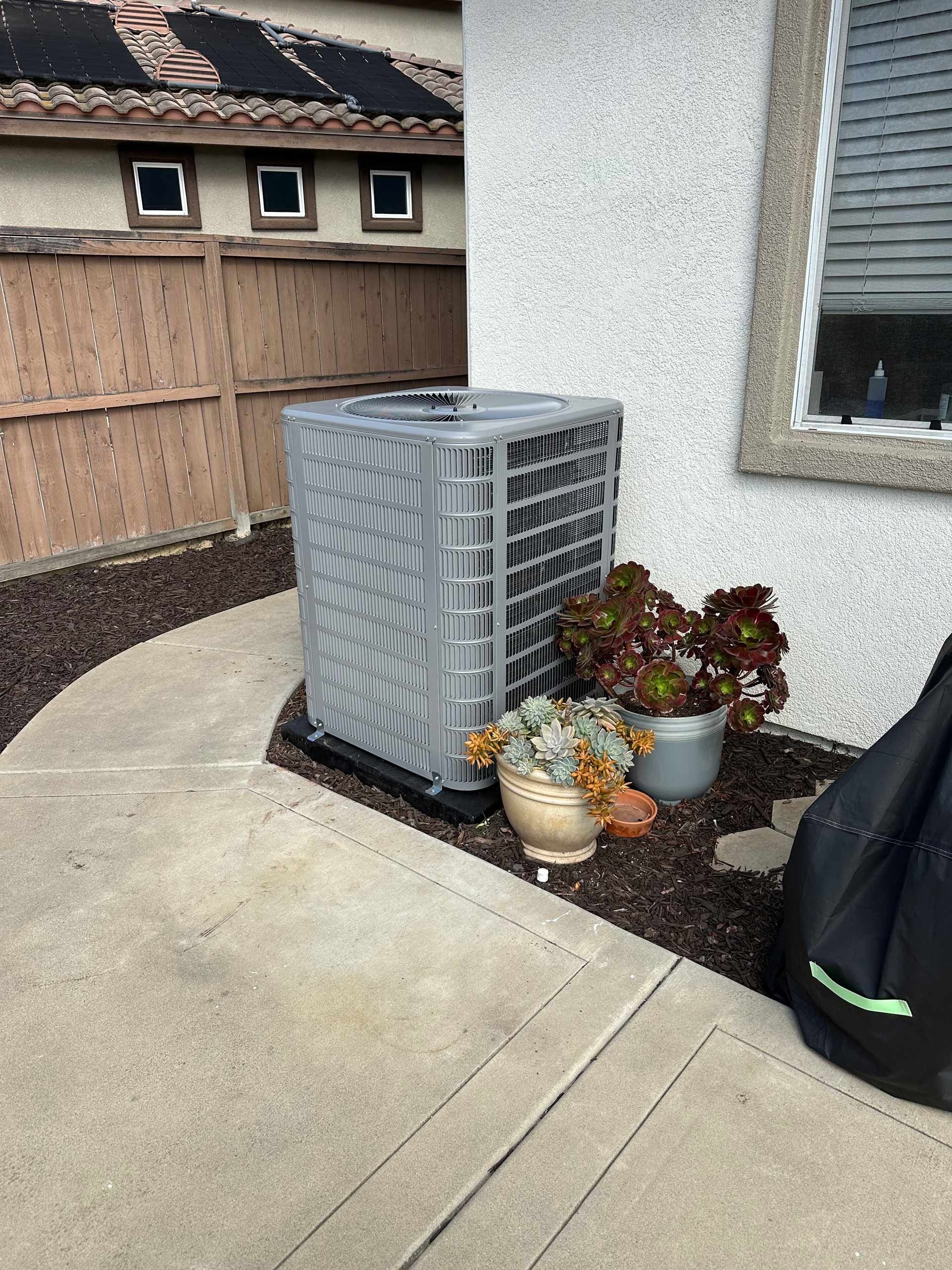 A gray air conditioner is sitting on a sidewalk next to a house.