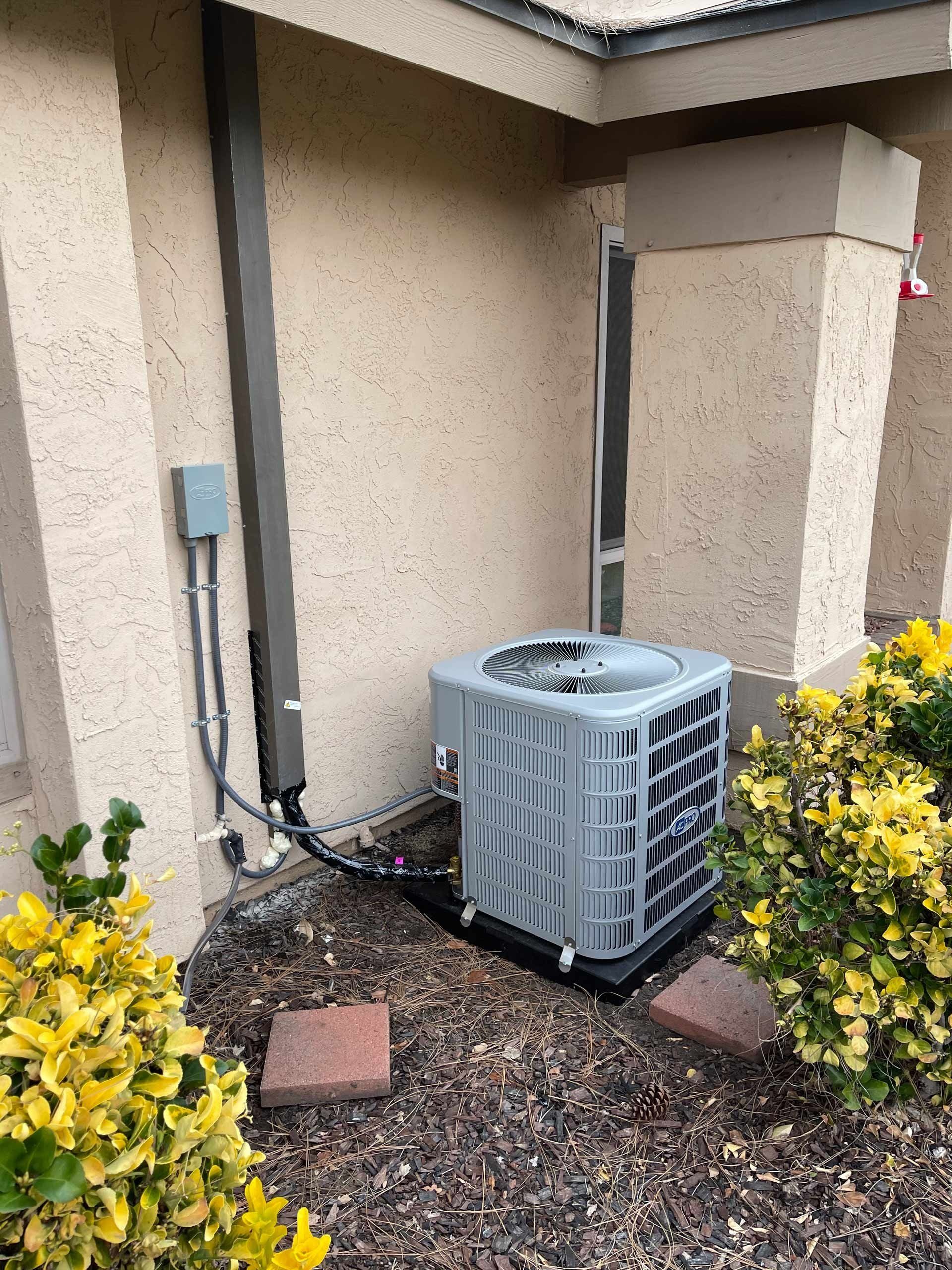 An air conditioner is sitting outside of a house next to a bush.