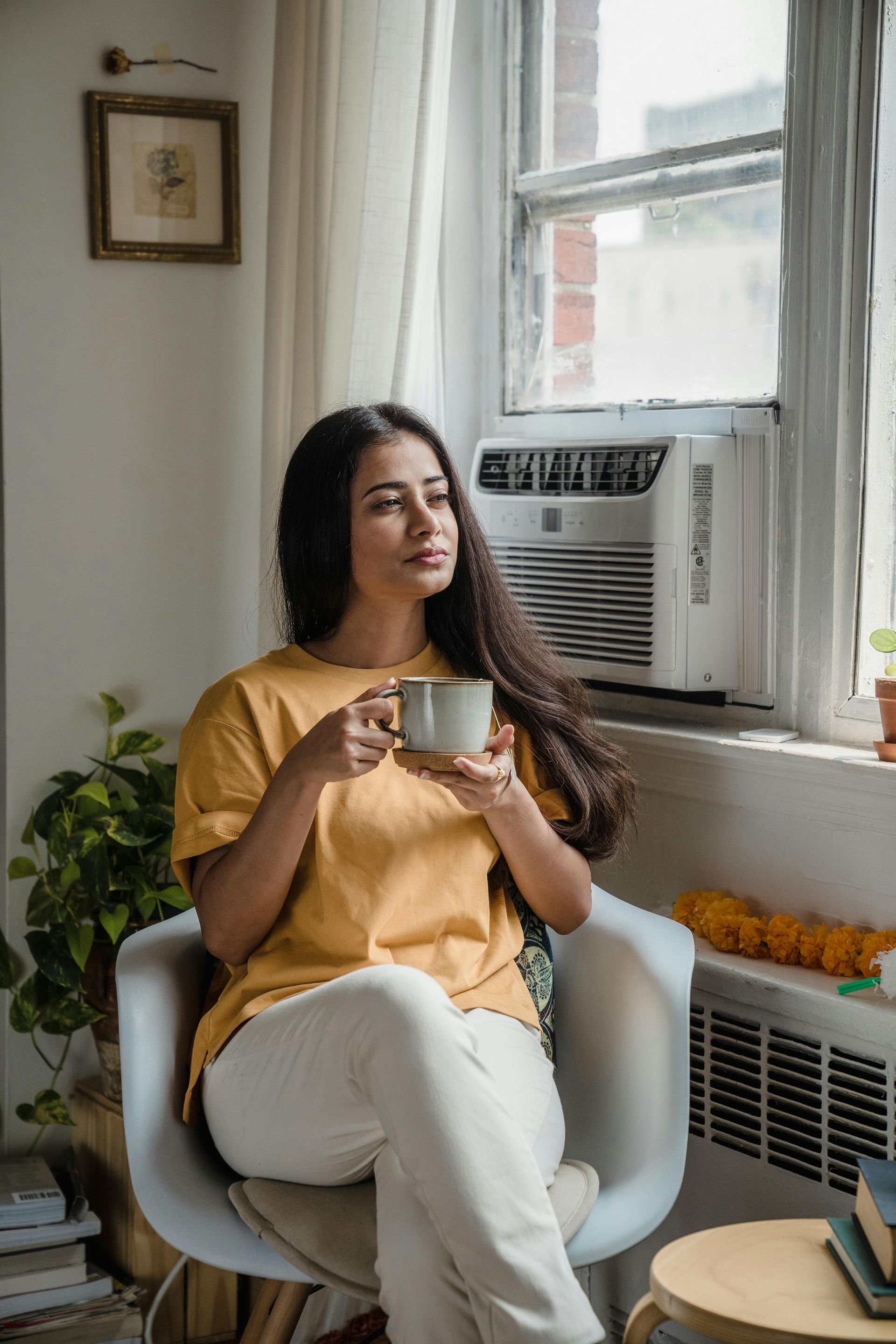 Woman in yellow shirt sits by window, holding mug, looking away.