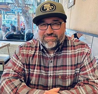 Man in a flannel shirt and 49ers cap smiles, wearing glasses. Seated at a table in an outdoor cafe.