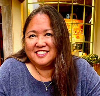 Smiling woman with long brown hair, wearing a blue sweater, seated in front of a window.