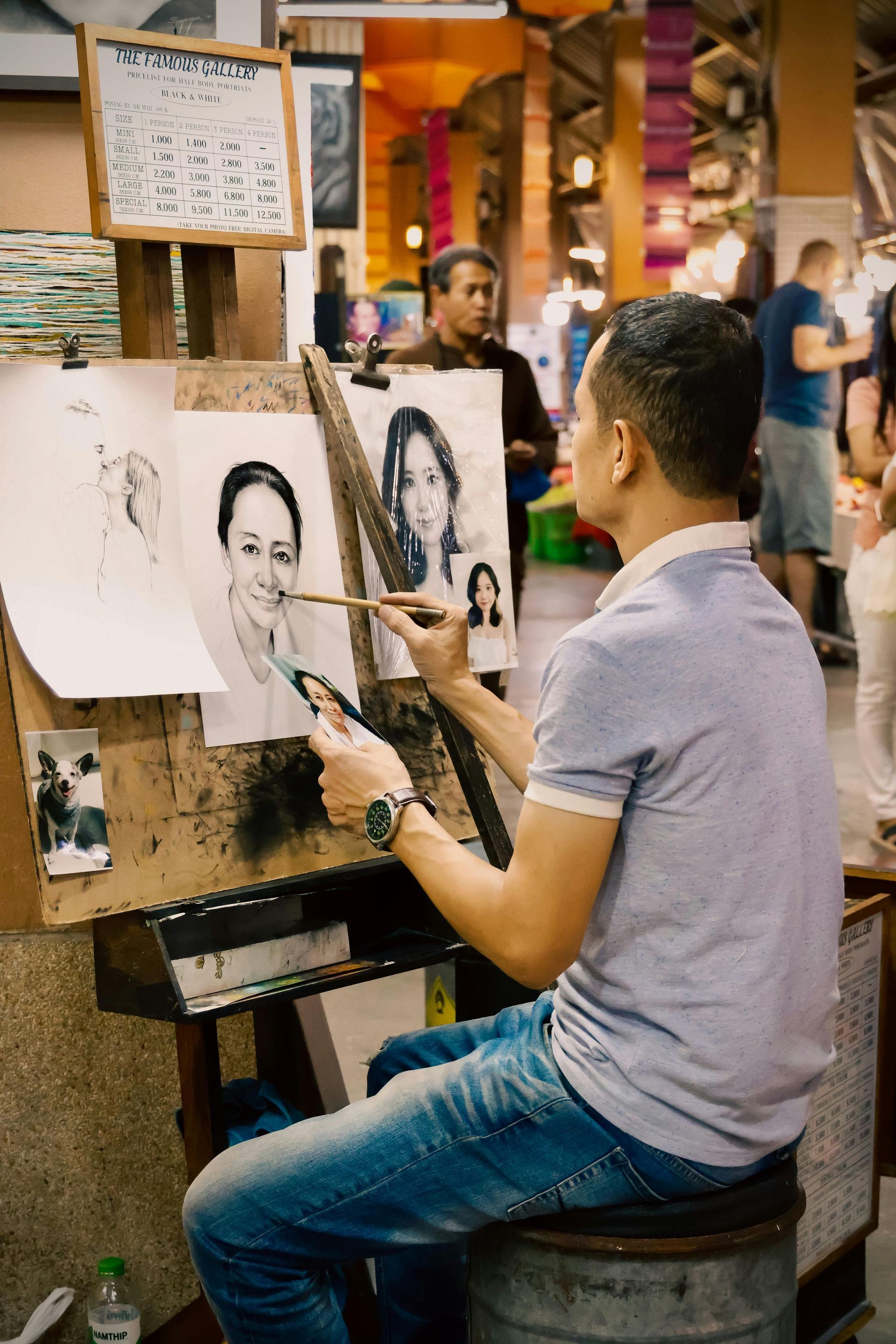 Artist at work sketching portraits in a busy market. He wears jeans and a light blue shirt.