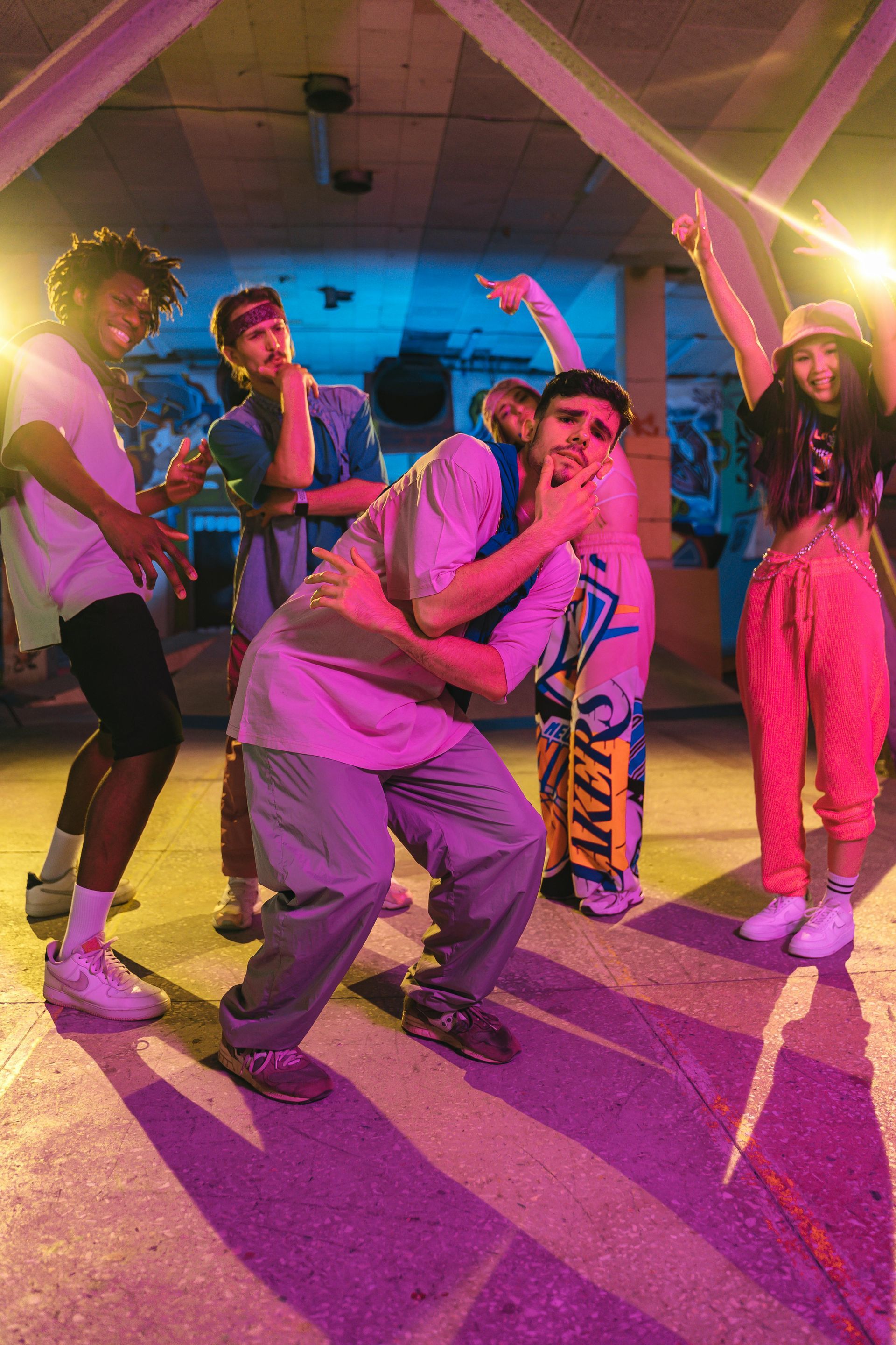 Group of people dancing in a brightly lit indoor space. One is embraced, others pose. Neon lights illuminate.