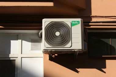 Outdoor air conditioner unit mounted on a building wall, with a fan grille and green label.