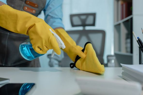 A person wearing yellow rubber gloves wipes a white office desk with a yellow cloth and a blue spray bottle.