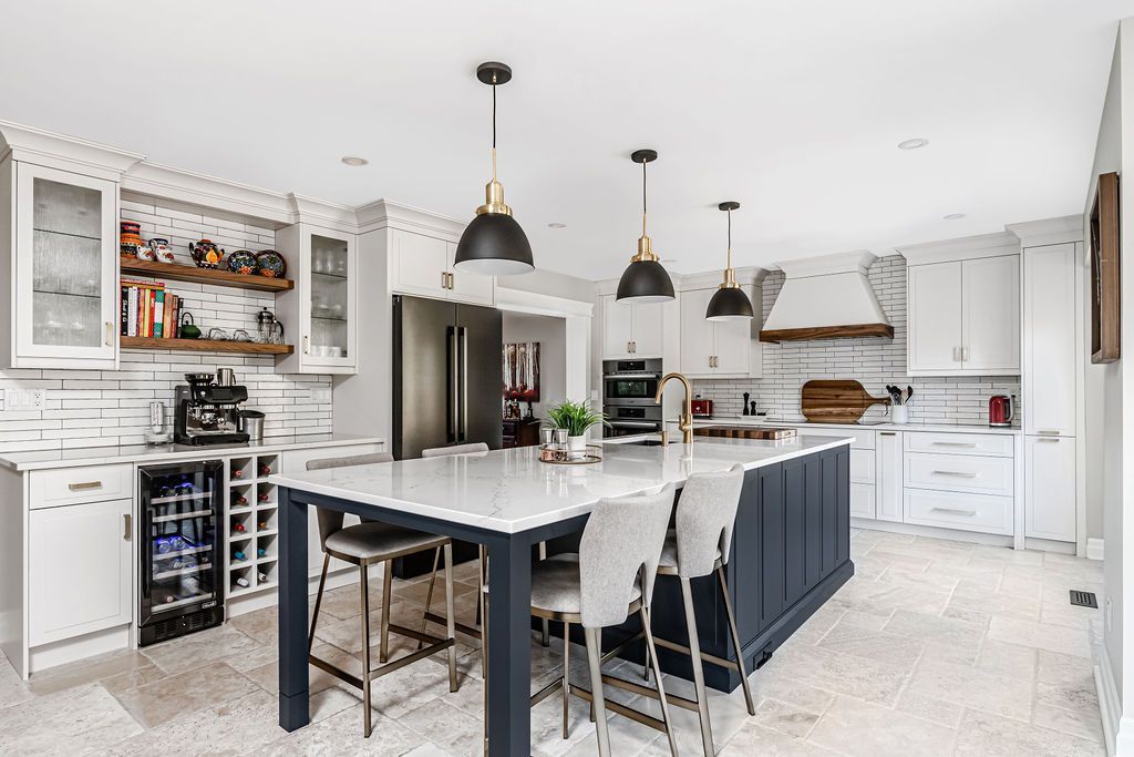 Modern white kitchen with dark blue island, pendant lights, and stainless steel appliances.