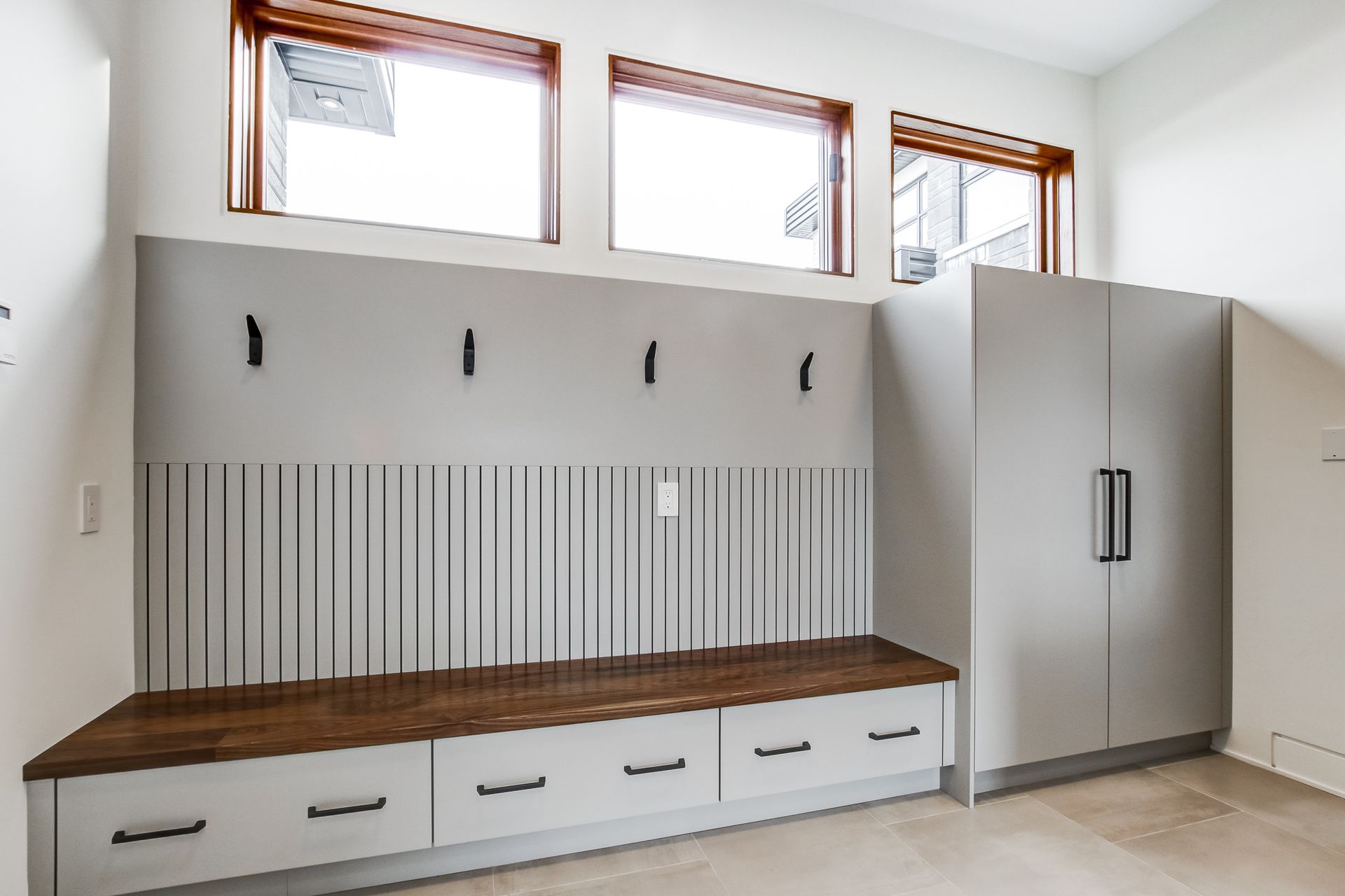 Mudroom with gray cabinets, wooden bench, coat hooks, and windows.