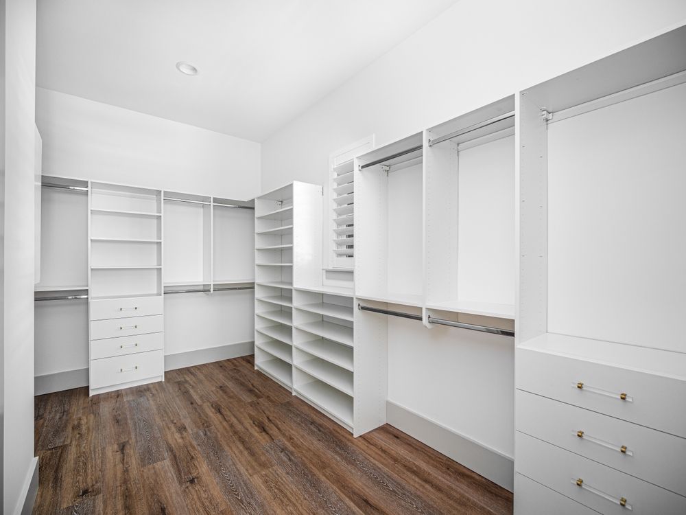 A walk-in closet featuring white modular shelving, hanging rods, and built-in drawers over dark wood-look flooring.