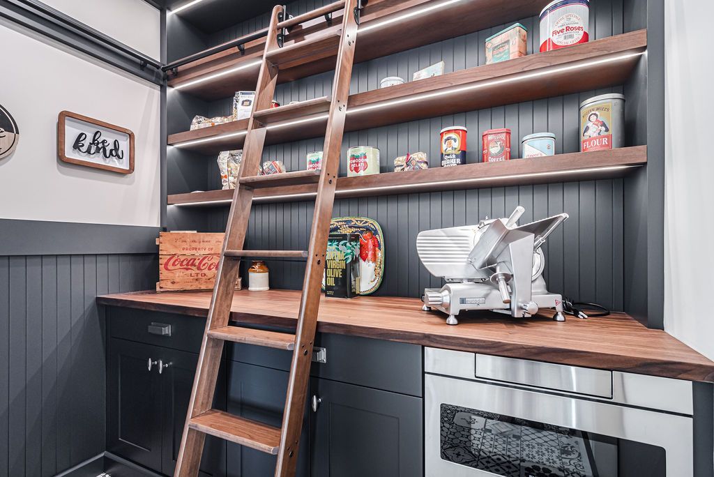 Pantry with wooden shelves, ladder, and a meat slicer. Dark cabinetry and countertop, vintage food cans on display.