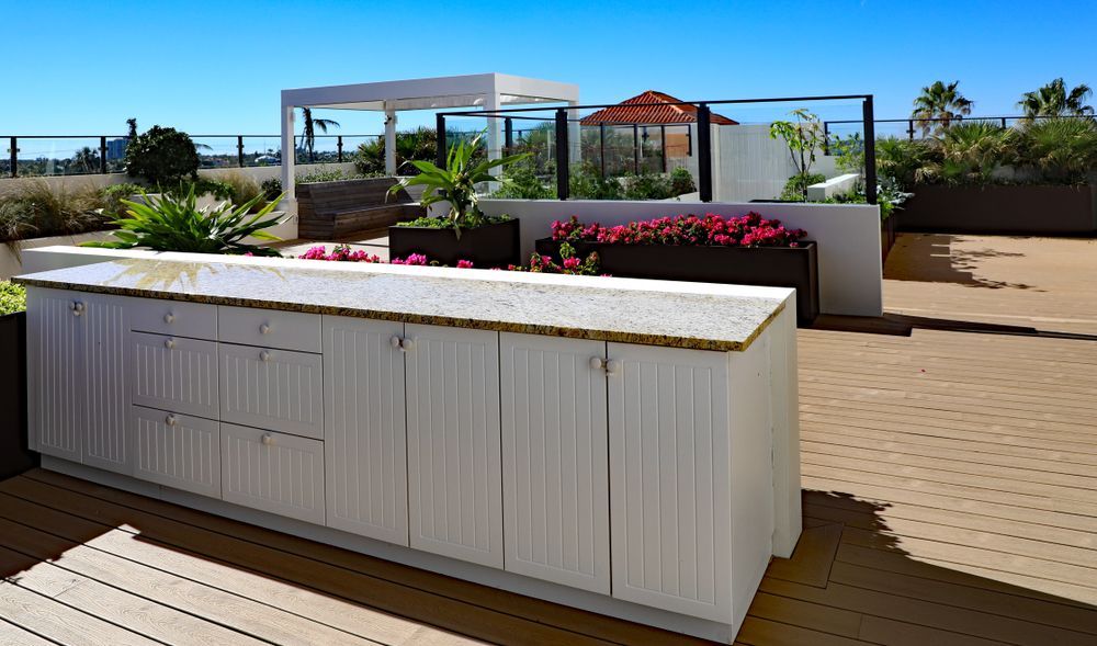 A white outdoor kitchen island with a stone countertop on a spacious wooden deck with planters under a clear blue sky.