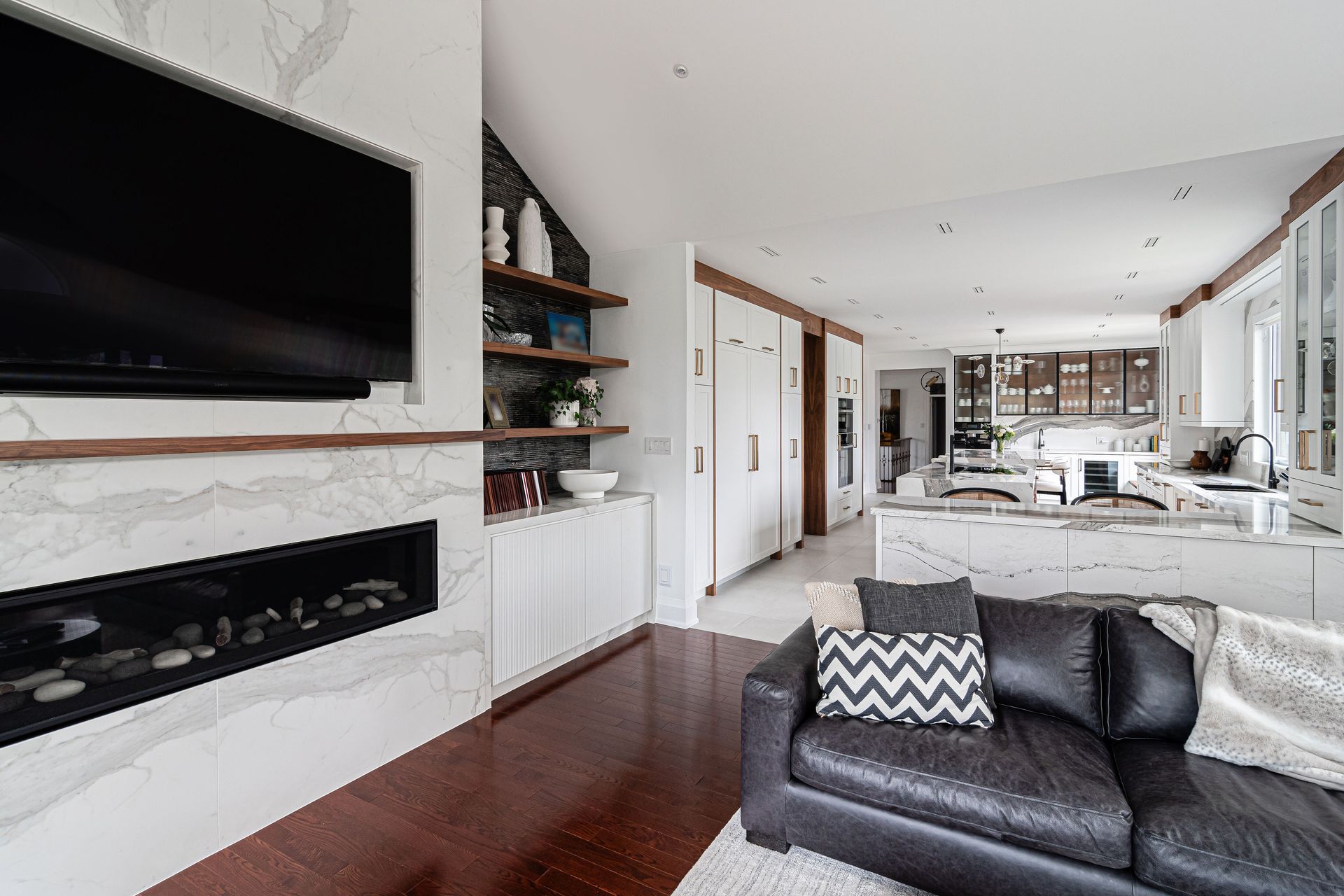 Living room with fireplace, TV, shelves, and leather sofa in front of a kitchen island.