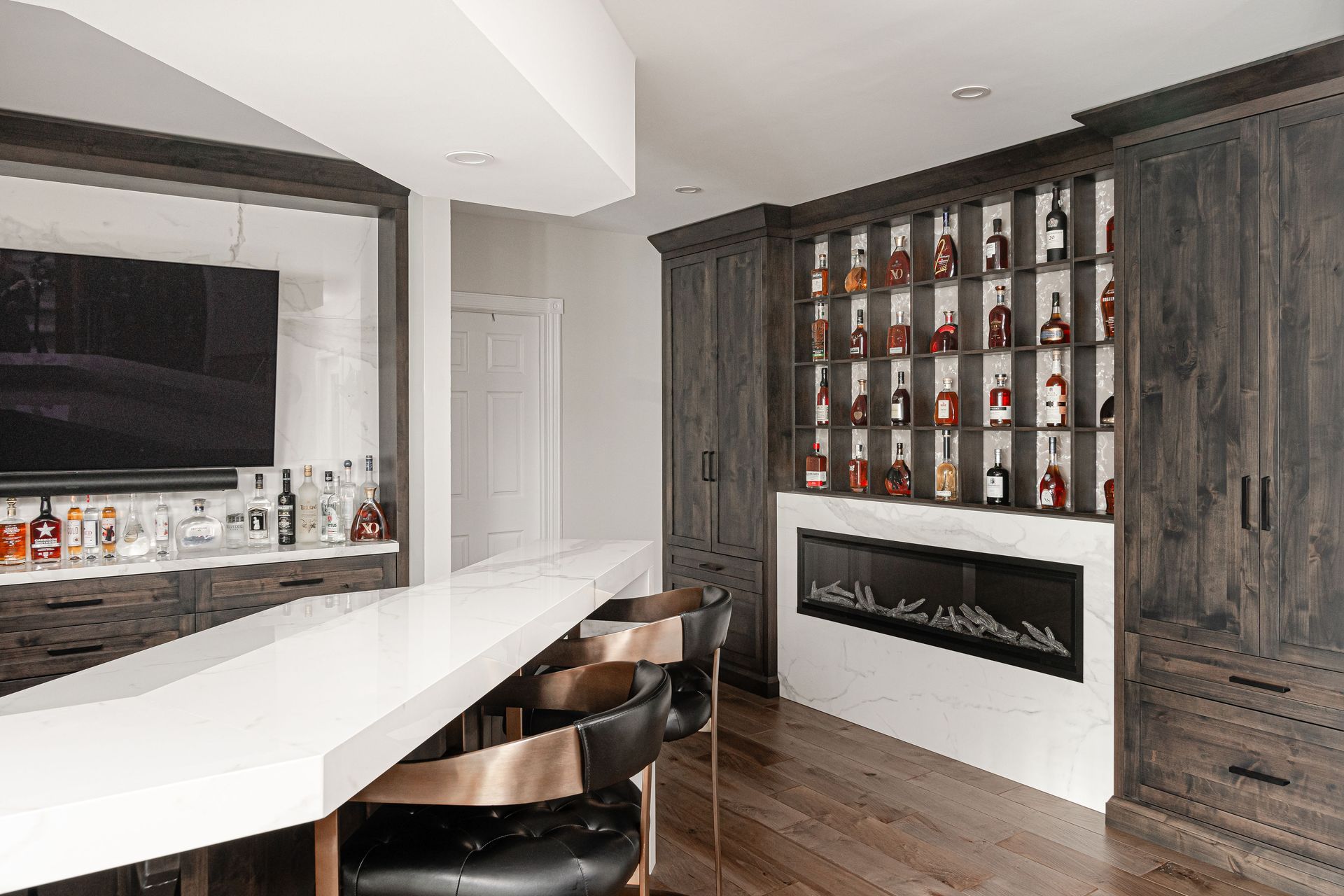 A modern basement bar with a white countertop, liquor display, fireplace, and black leather bar stools.