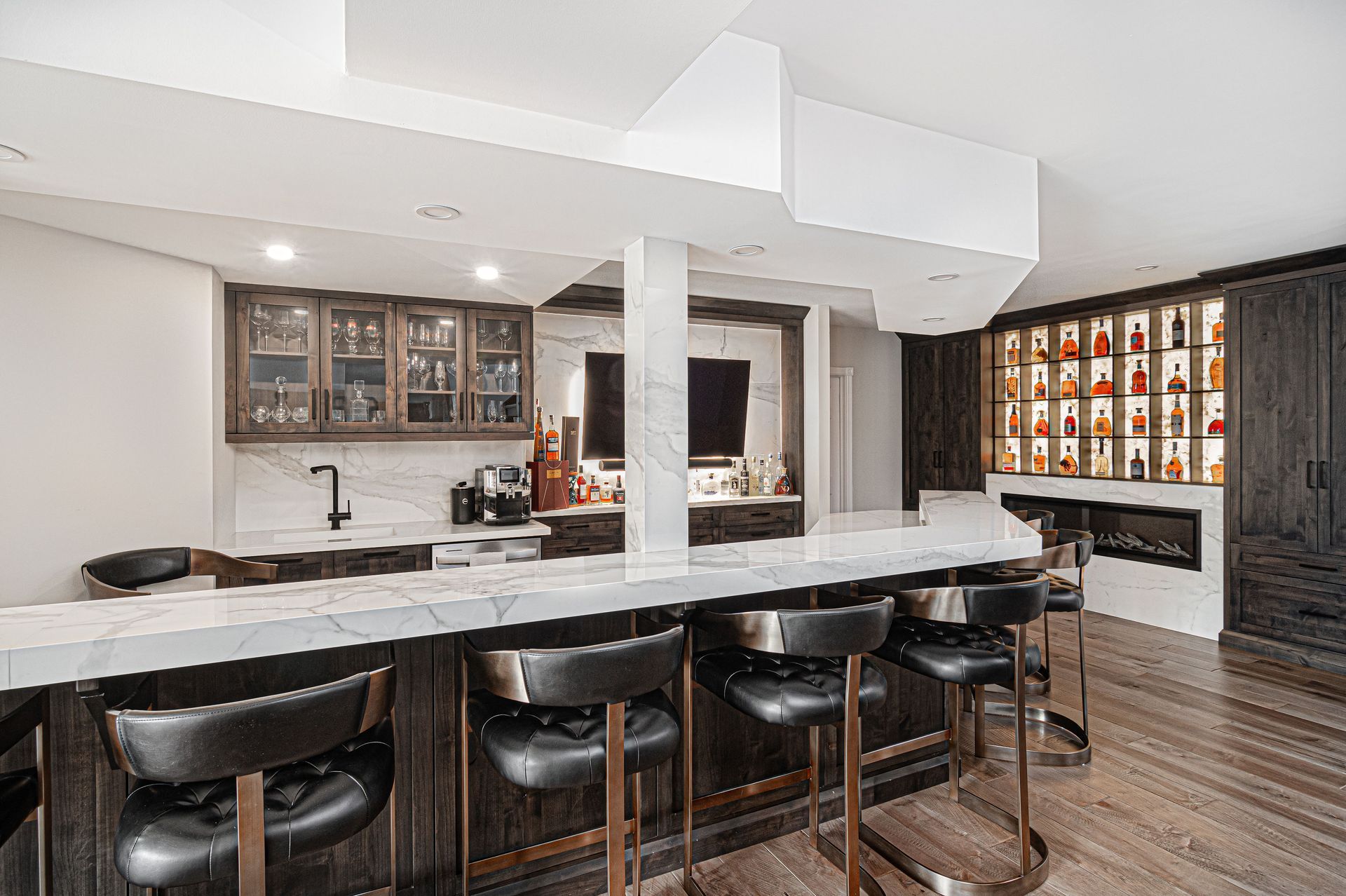 Interior view of a modern home bar with stools, cabinetry, and a fireplace. Dark wood and marble accents.